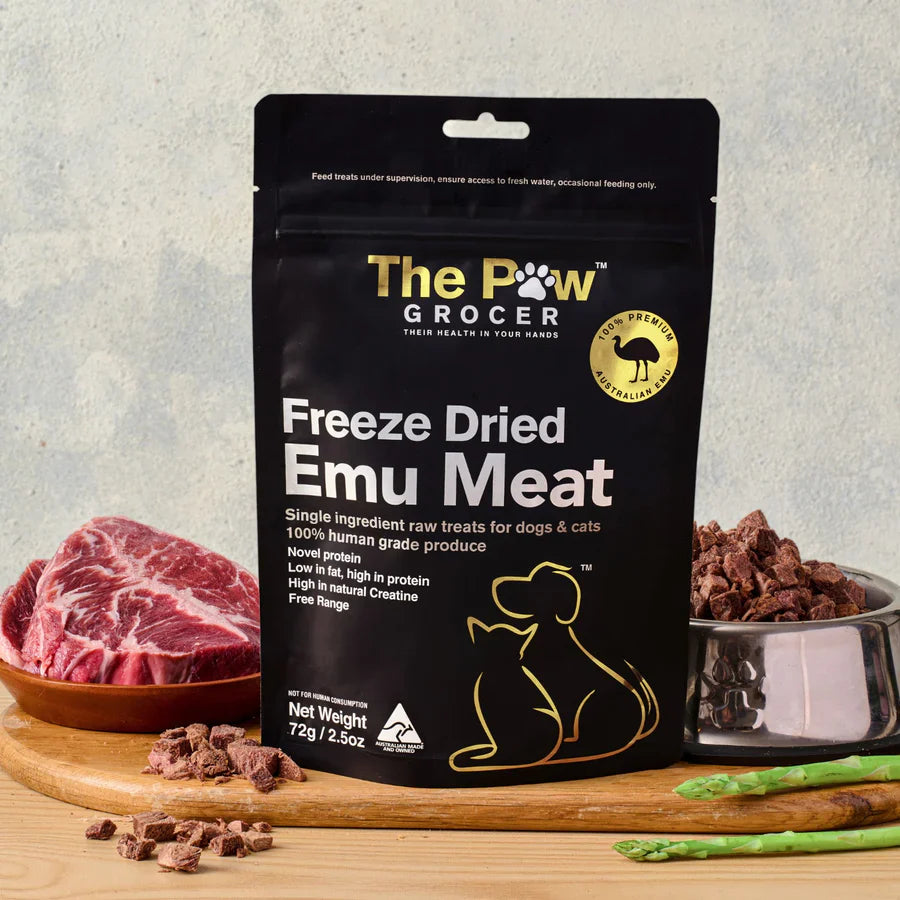 The Paw Grocer 72g Freeze-Dried Emu Meat treats displayed on a wooden board next to a cut of fresh raw meat and a silver bowl filled with the crunchy, hypoallergenic cubes.