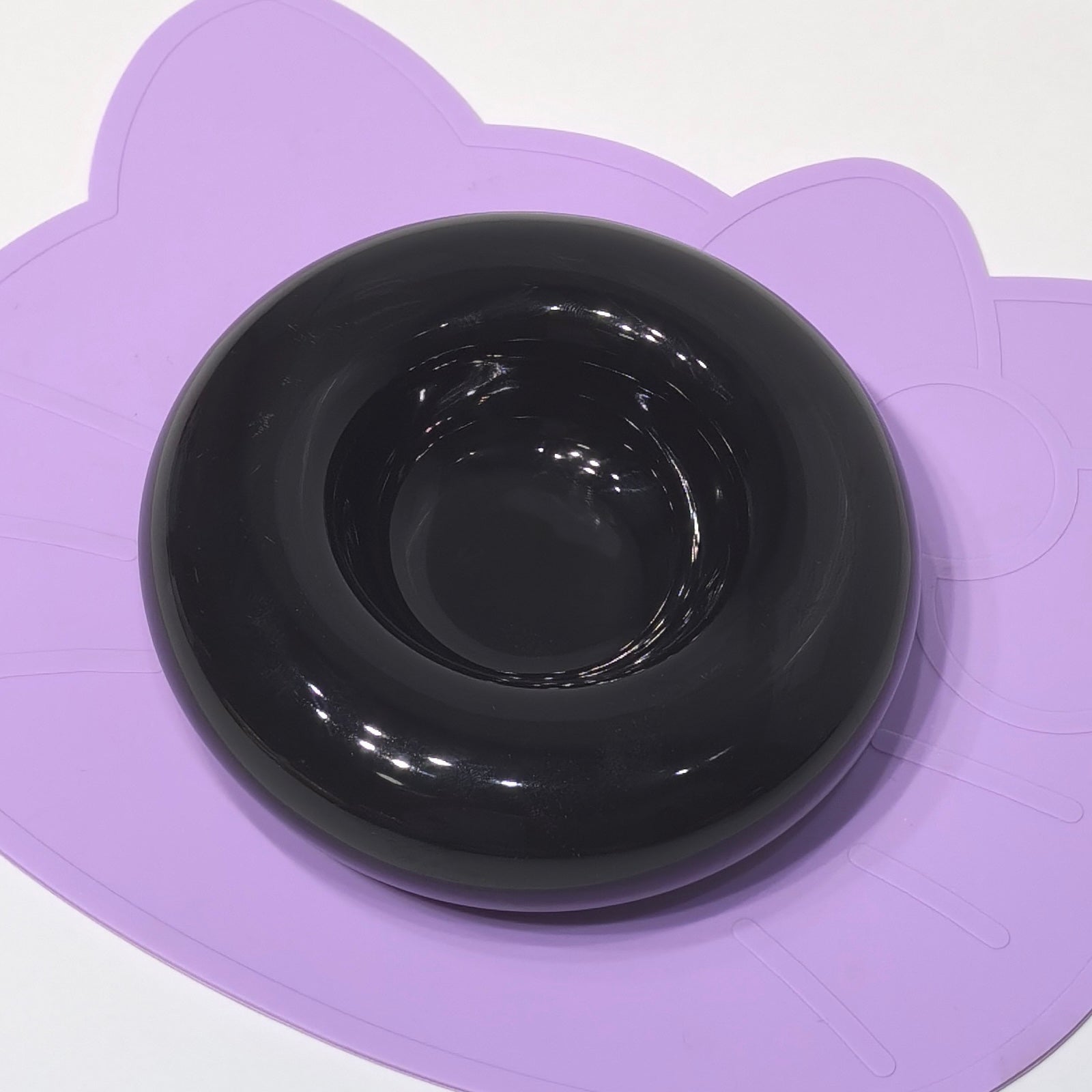 A high-gloss black ceramic bagel-shaped pet bowl placed on a purple cat-shaped silicone feeding mat, showing its smooth reflective surface and deep center well.