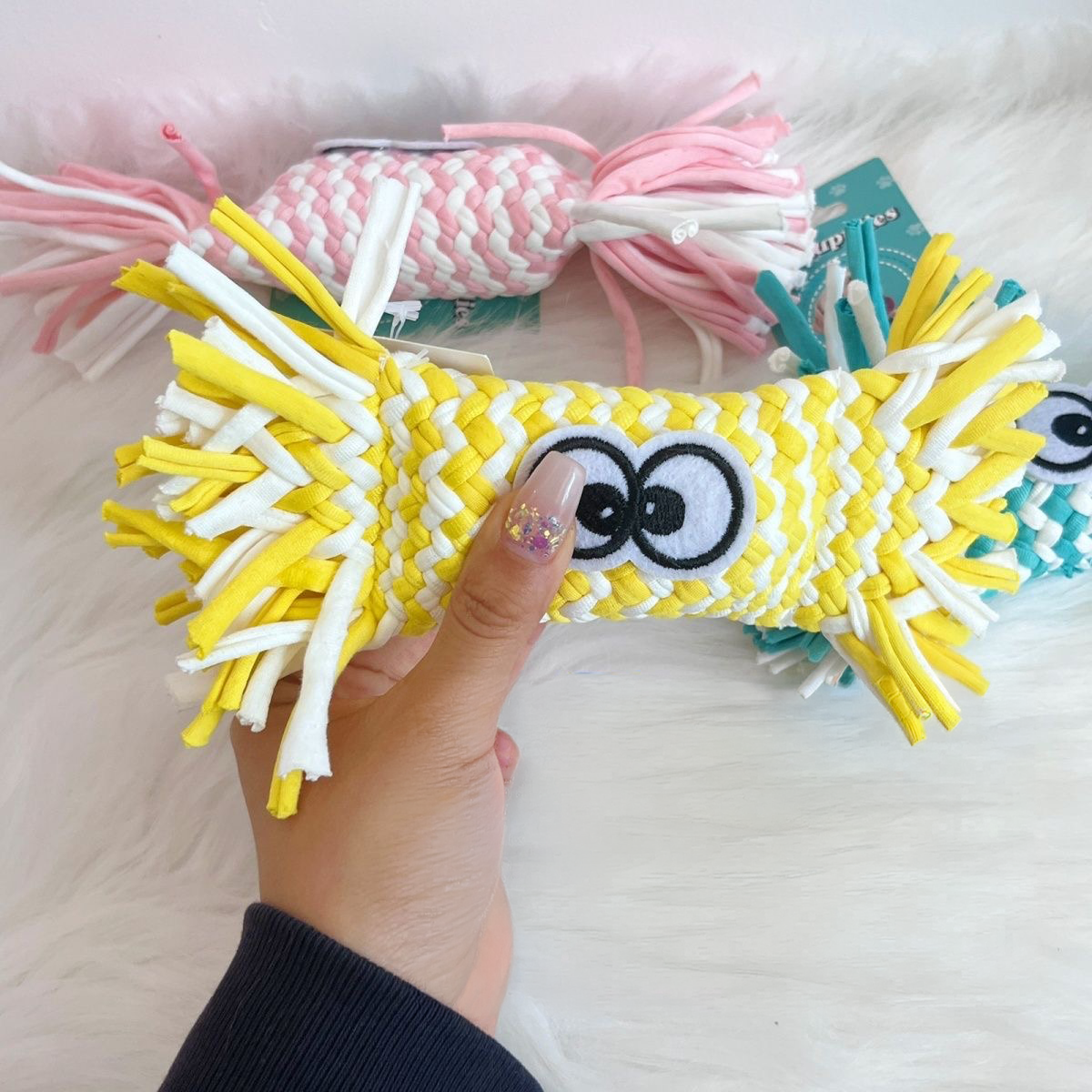 A hand holding a bright yellow woven candy dog toy, showcasing the thick braided stretch fabric strips and the funny embroidered cartoon eyes, highlighting its durable construction.