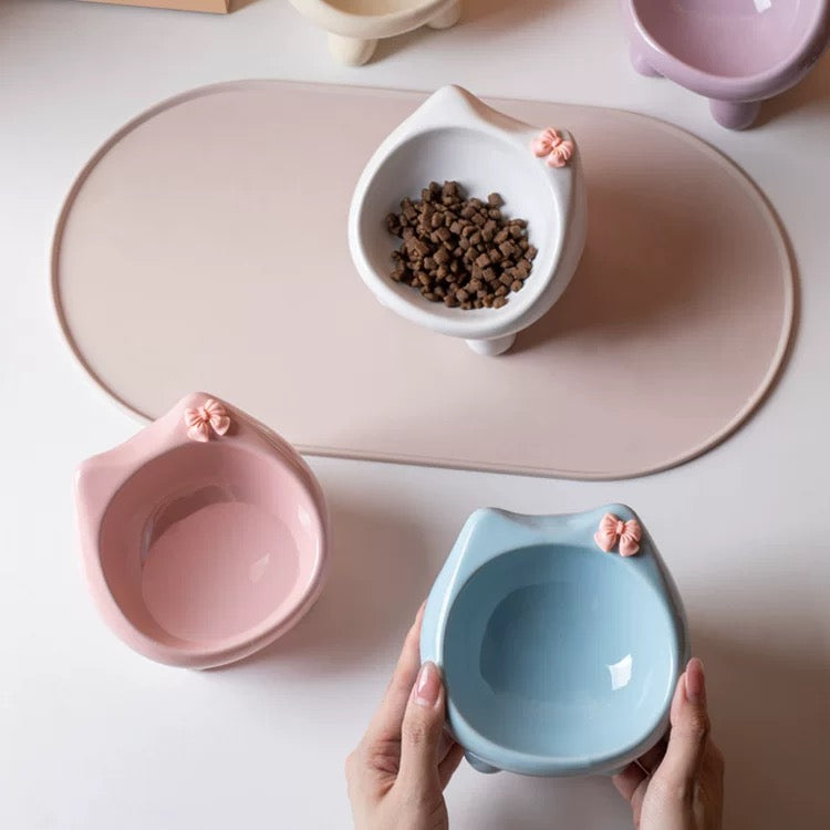 A pair of hands holding a light blue cat-ear ceramic pet bowl above a pink mat, highlighting the smooth anti-black chin interior and cute bow detail.
