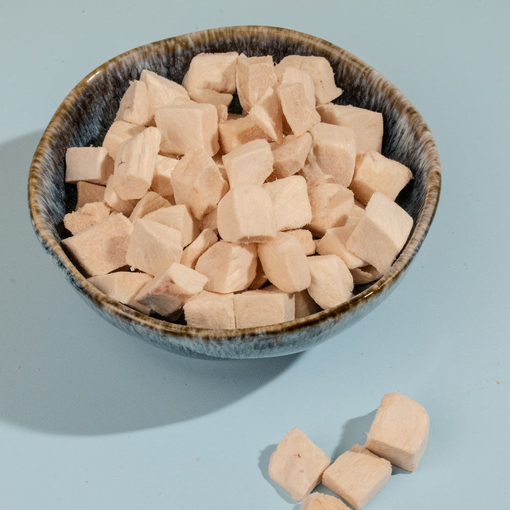 A dark ceramic bowl filled with uniform freeze-dried chicken cubes on a light blue background, showcasing a classic and highly palatable reward for cats and dogs.