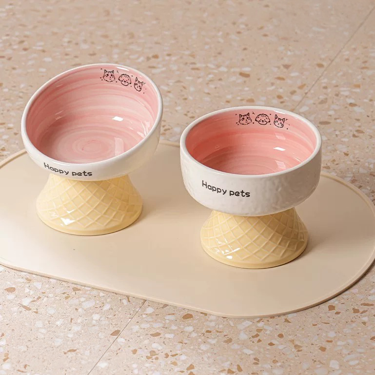 Two pink ceramic pet bowls styled on a beige mat. The high-foot design reduces neck strain for adult cats