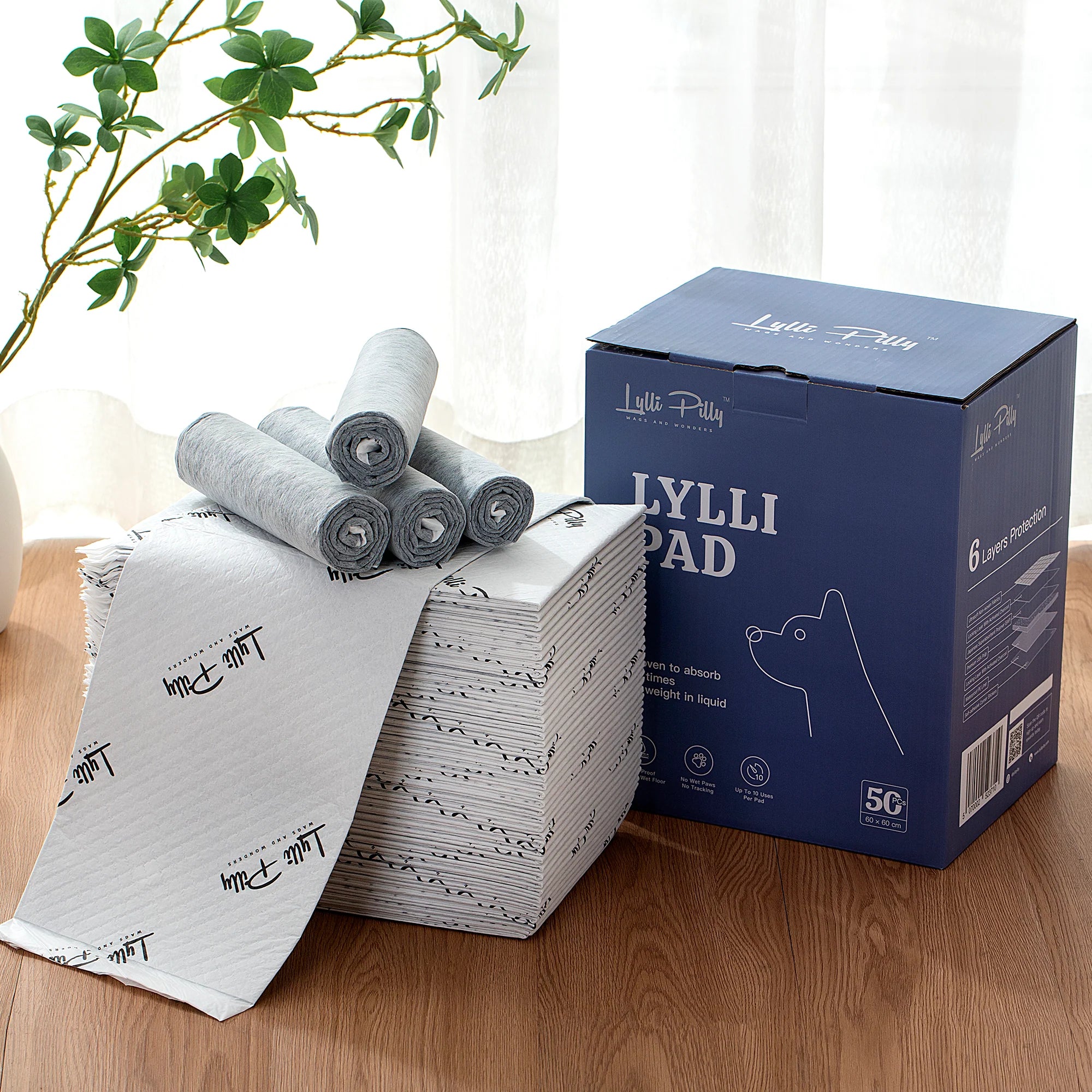 A stack of LYLLI PILLY premium pet training pads next to their blue 50-piece packaging box, featuring rolled-up grey pads on top.