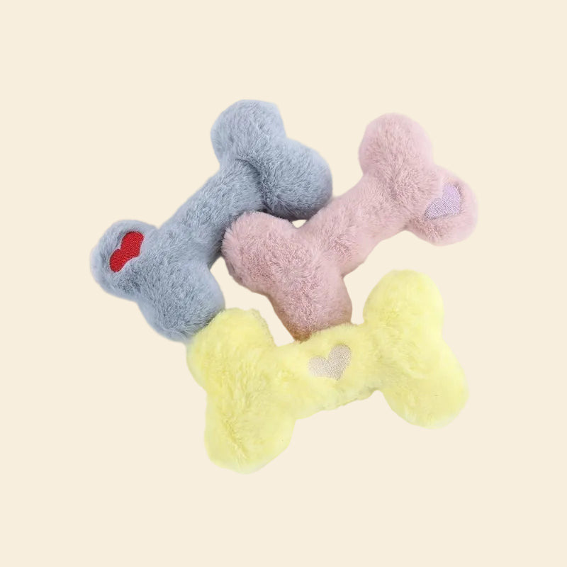 Three pastel-colored plush bone dog toys—sky blue, soft pink, and lemon yellow—stacked together on a pale background. Each fluffy bone features a small, cute embroidered heart in the corner.