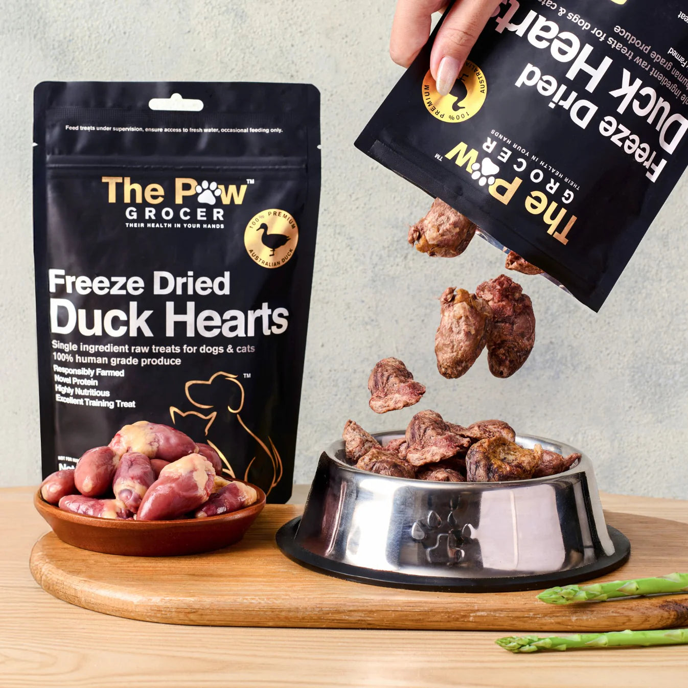 A hand pouring 100% pure freeze-dried duck hearts from the pouch into a silver pet bowl, showcasing the raw, iron-boosted muscle meat texture that fortifies long-term health.