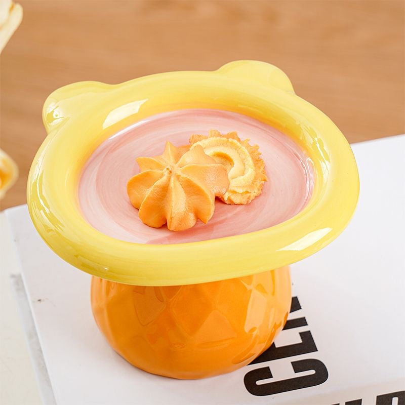 Top-down view of the yellow and pink Teddy Bear Elevated Ceramic Pet Bowl holding pet treats. The wide, shallow dish sits on a textured orange geometric pedestal base.