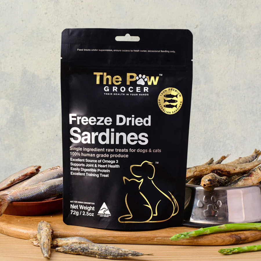 The Paw Grocer Freeze Dried Sardines (72g) product bag front, displayed in a rustic kitchen setting on a wooden board. It features a bowl of raw whole sardines, a bowl of dried sardine treats, scattered dried fish, and asparagus spears. The bag details: single ingredient, 100% human grade produce, Omega-3 source, and dog/cat illustrations.