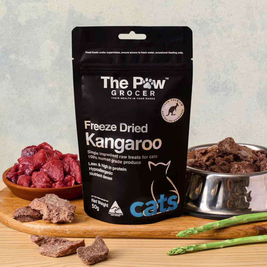 The Paw Grocer Freeze-Dried Kangaroo cat treats packaging on a wooden board between bowls of raw meat and dried treats. Highlighting transparency from raw to treat.