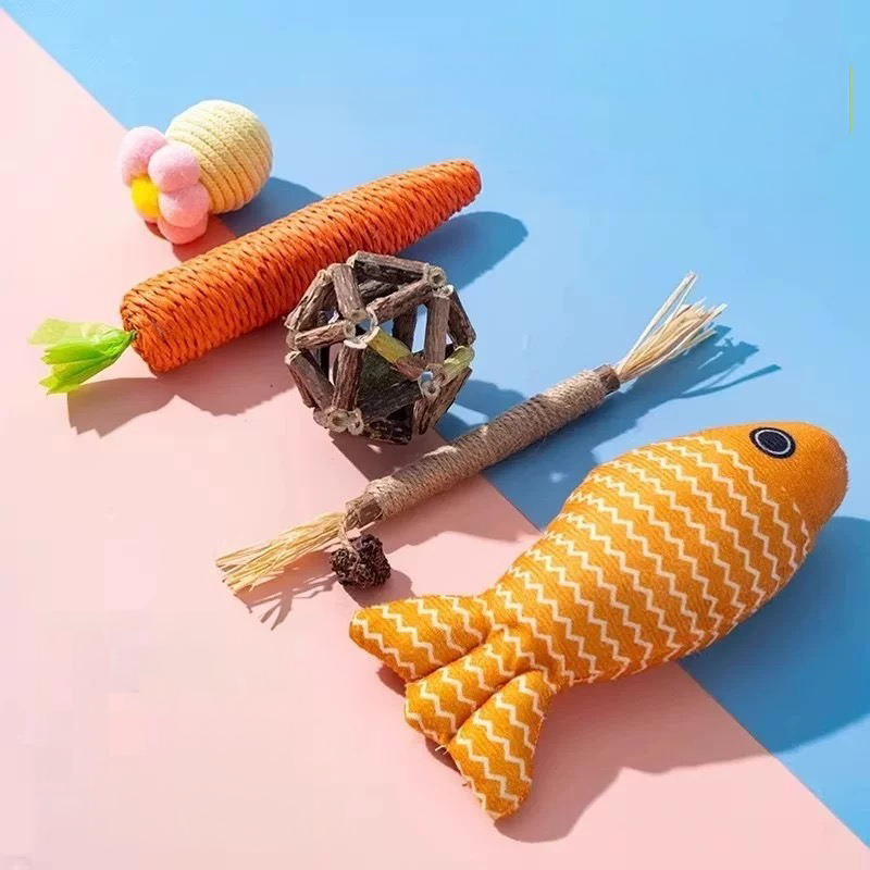 A collection of five premium feline enrichment toys, including a hemp carrot, silvervine cage ball, teething stick, yellow catnip fish, and pastel rattle ball, arranged diagonally on a pink and blue background.