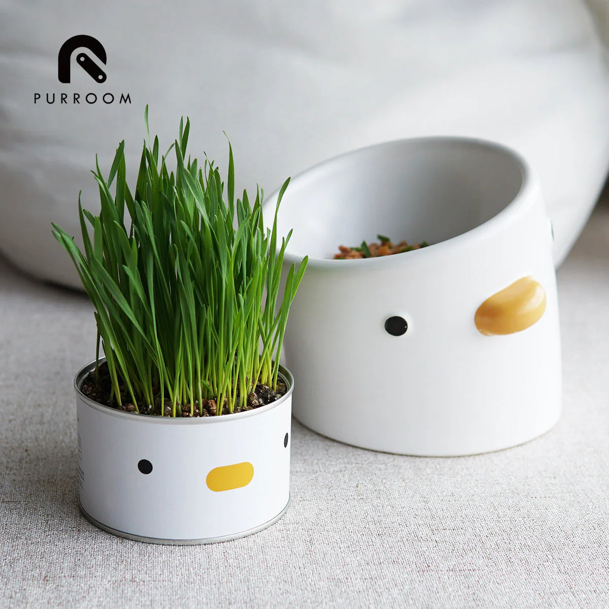 Small white planter with green grass next to a larger white bowl with a chick design on a light surface.