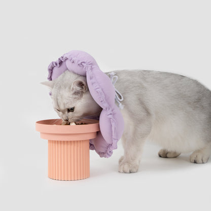 A cat easily eating from an elevated pink bowl while wearing the soft purple HiDREAM recovery collar, proving it does not block access to food.