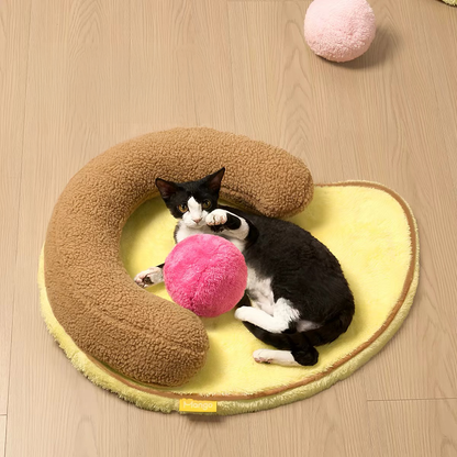 A cute tuxedo cat resting comfortably inside the U-shaped bolster of the Mango Pet Bed while playfully batting at the attached pink fluffy play ball.