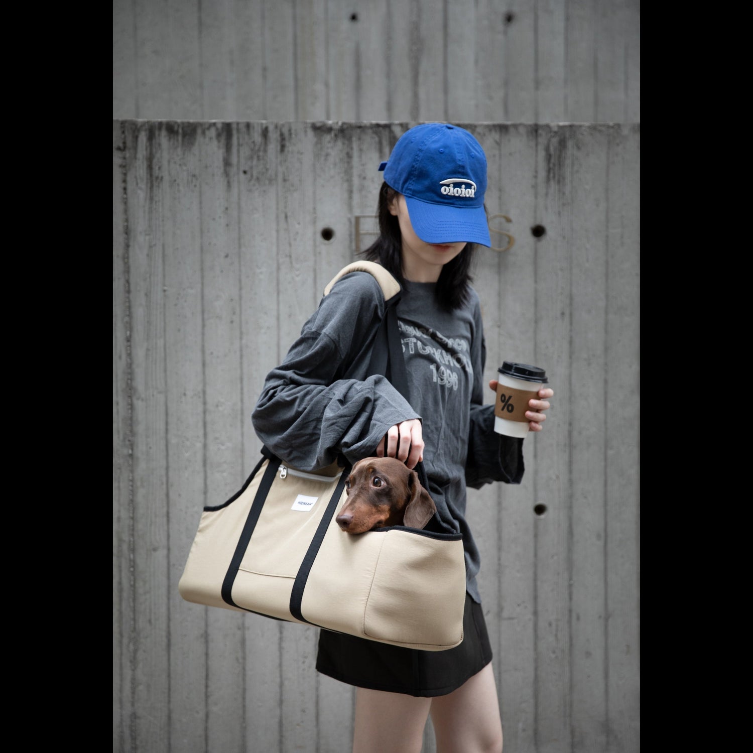 Person holding a brown tote bag with a small dog peeking out, wearing a dark jacket and holding a coffee cup.