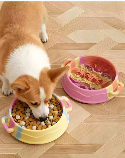 A cute Corgi dog actively eating dry kibble from a yellow and blue silicone slow feeder bowl on a wooden floor, with a second pink bowl filled with wet food placed nearby to demonstrate healthy eating habits.
