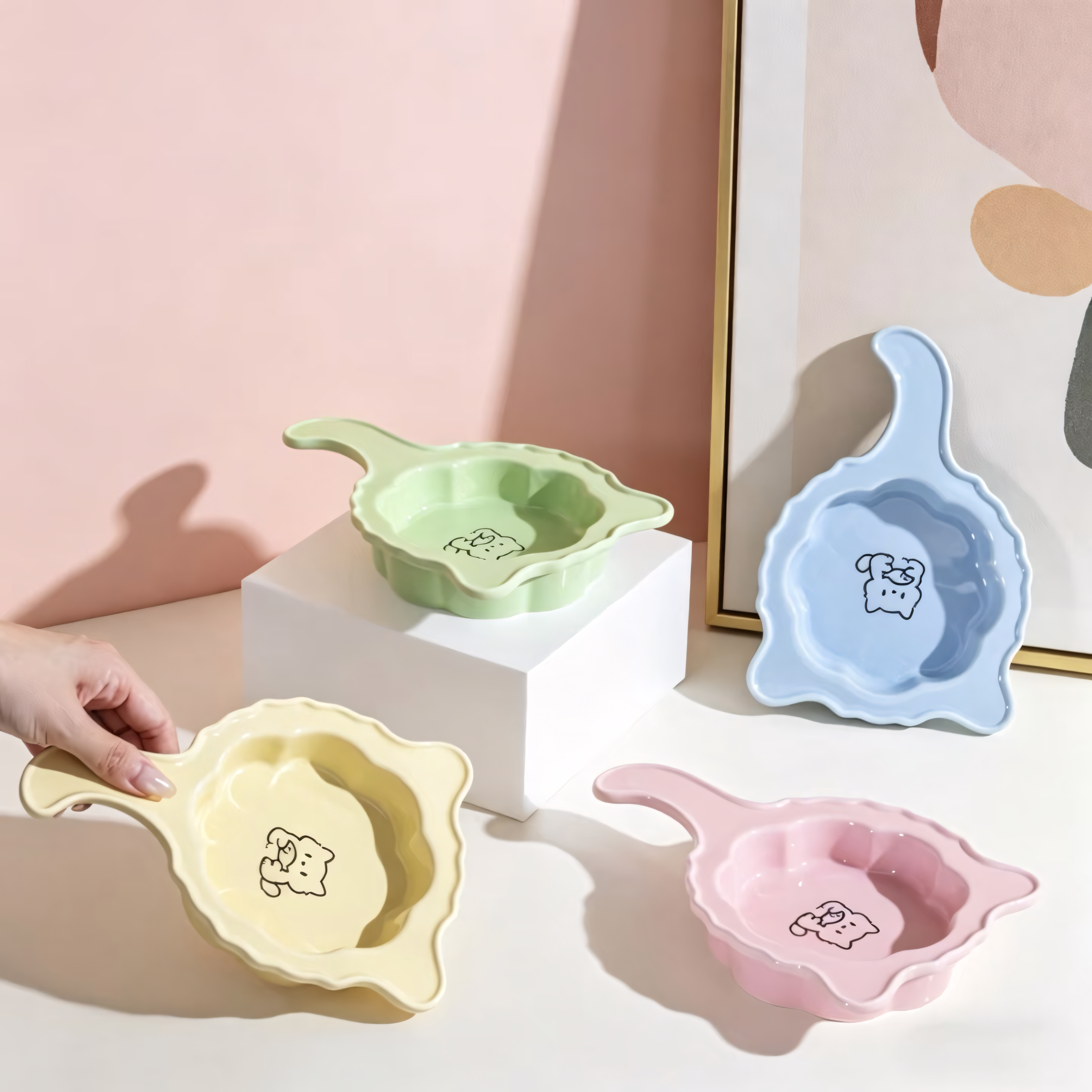 A hand effortlessly lifting the Butter Yellow Crooked Tail Ceramic Pet Plate by its ergonomic handle. Also displaying the Soft Pink, Baby Blue, and Mint Green plates.