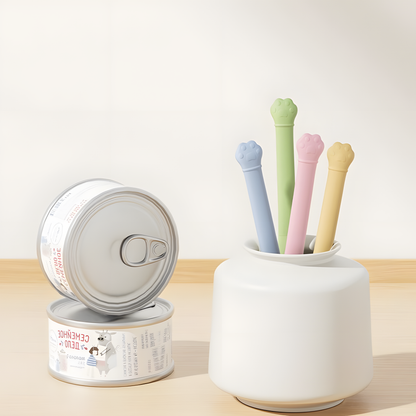 Four pastel-colored silicone pet food spatulas with cute paw print handles stored neatly in a white ceramic vase next to stacked pet food cans.