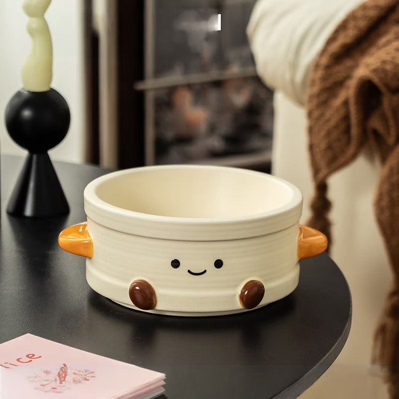 A lifestyle shot of the cute Dim Sum Steamer ceramic pet bowl resting on a black table. Features a therapeutic smiley face, orange side handles for easy lifting, and overlaid dimensions showing a 15cm inner diameter.