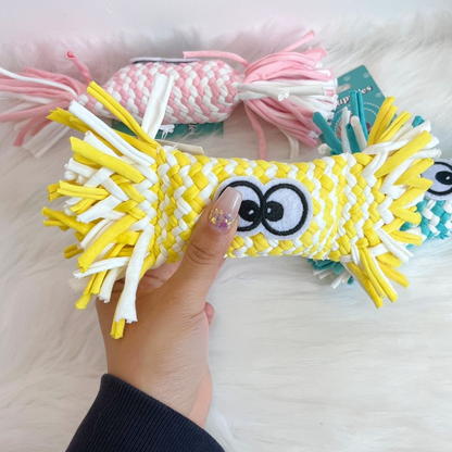 A hand holding a bright yellow woven candy dog toy, showcasing the thick braided stretch fabric strips and the funny embroidered cartoon eyes, highlighting its durable construction.