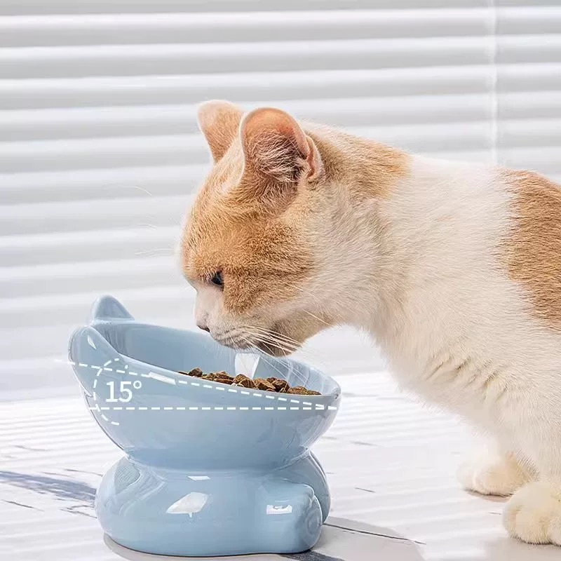 An orange and white cat comfortably eating from the light blue elevated ceramic bowl, with a graphic showing the ergonomic 15-degree tilt designed to protect the cervical spine.