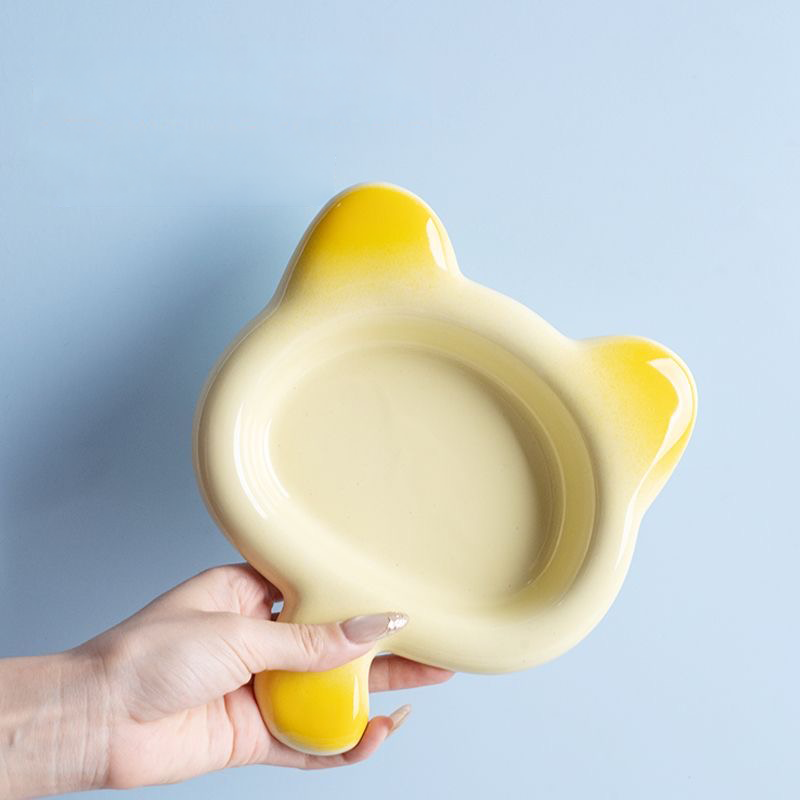 A hand comfortably lifting the yellow Chubby Cat ceramic plate by its clever tail handle. An ergonomic, mess-free design for serving wet pet food.