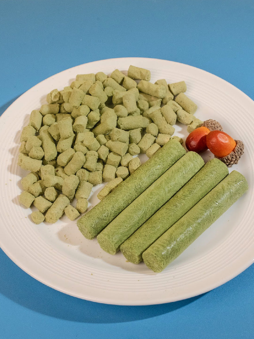 A white plate displaying whole green freeze-dried cat grass sticks alongside smaller, bite-sized broken pieces, showcasing the high-protein, crunchy texture designed to aid feline digestion and dental health.