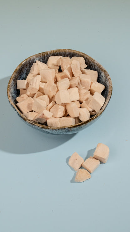 A dark ceramic bowl filled with uniform freeze-dried chicken cubes on a light blue background, showcasing a classic and highly palatable reward for cats and dogs.