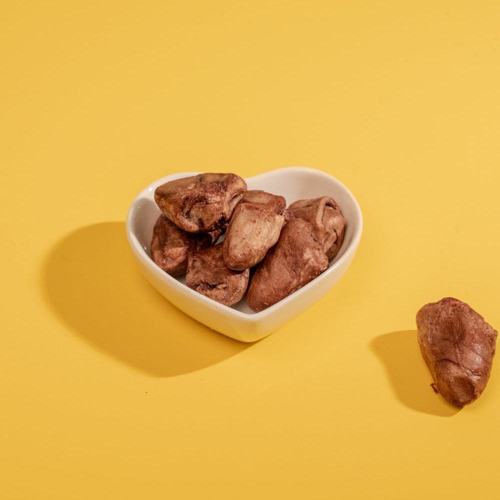 Premium freeze-dried chicken heart treats beautifully presented in a small white heart-shaped ceramic bowl against a vibrant yellow background, illustrating a high-value, lovable reward for pets.