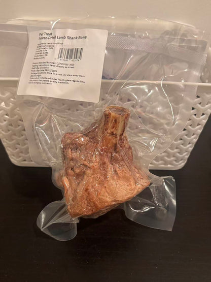 A real-world photo of a large freeze-dried lamb shank bone dog chew safely sealed in a clear vacuum bag. The attached label details the single-ingredient purity, guaranteed analysis, and safety warnings.