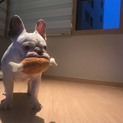 A white French Bulldog proudly carrying a large plush chicken leg toy in its mouth, showing the realistic drumstick design and perfect size for medium dogs.