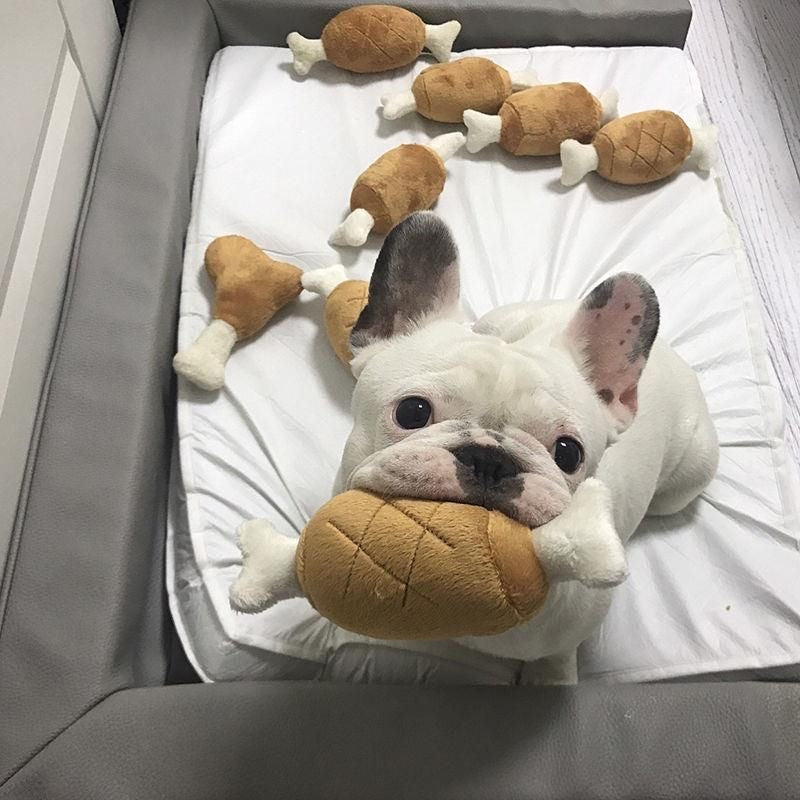 Looking down at a happy French Bulldog in its bed surrounded by several plush chicken legs, looking up excitedly with a toy in its mouth.