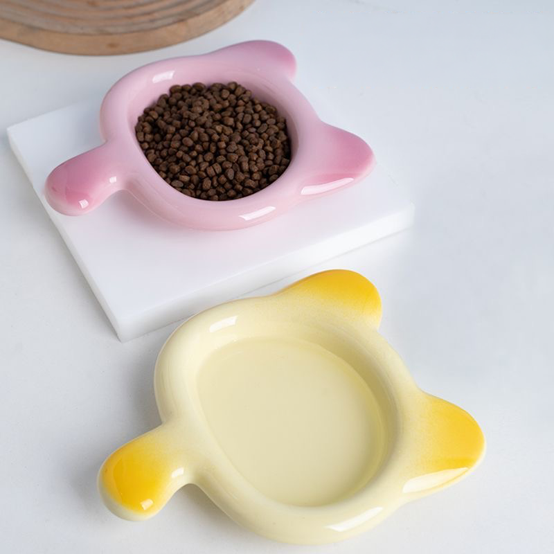 A pastel pink Chubby Cat Ceramic Pet Plate filled with dry kibble, sitting next to a yellow gradient plate. Showcasing the 17cm wide, whisker-friendly shallow basin for stress-free eating.