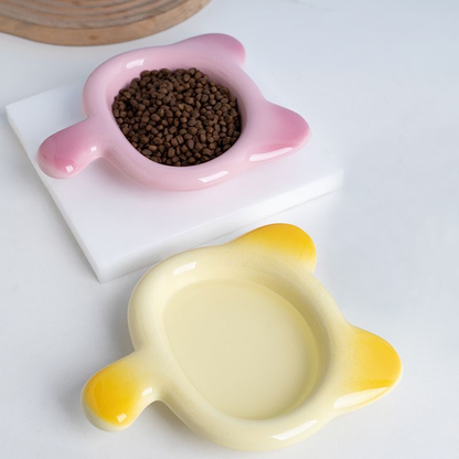 A pastel pink Chubby Cat Ceramic Pet Plate filled with dry kibble, sitting next to a yellow gradient plate. Showcasing the 17cm wide, whisker-friendly shallow basin for stress-free eating.