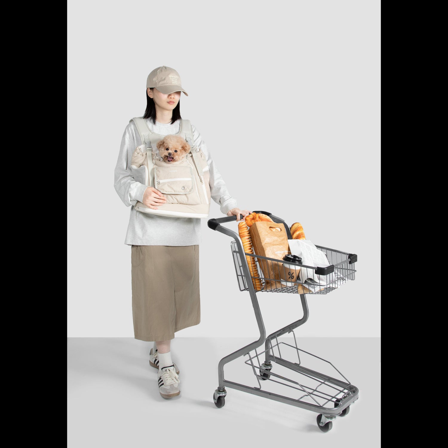 A woman easily pushing a grocery cart while comfortably carrying her small poodle in a beige, hands-free front pet carrier backpack.
