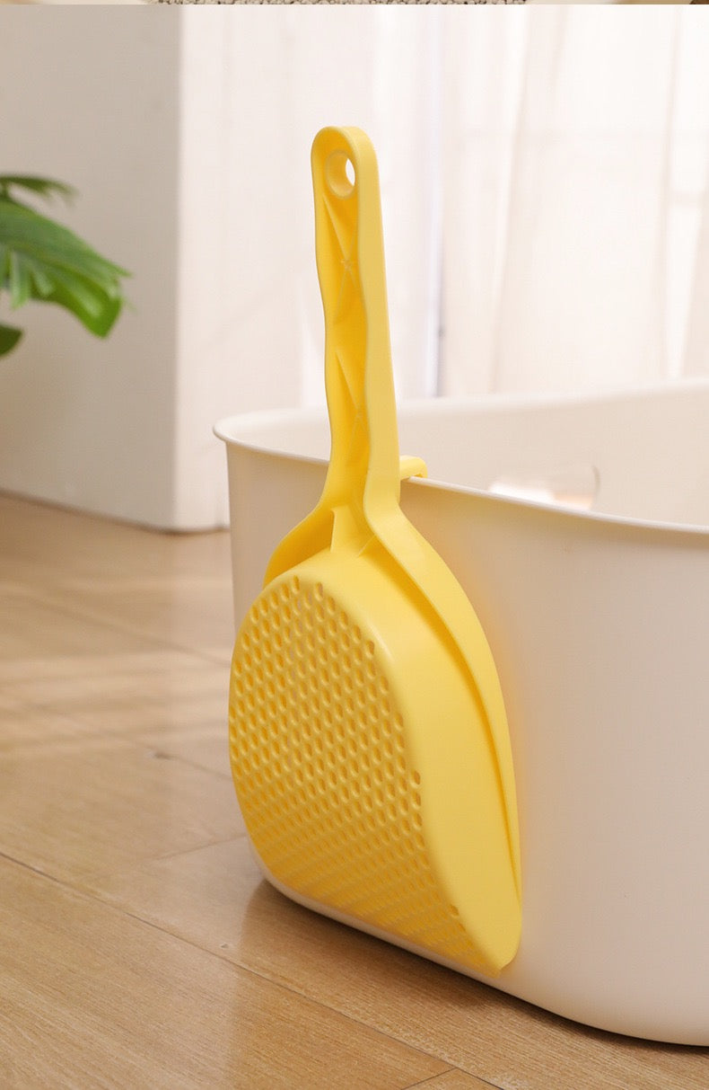 The yellow cat litter scoop conveniently hanging on the edge of a white litter box using its built-in back clip for hygienic, off-the-floor storage.
