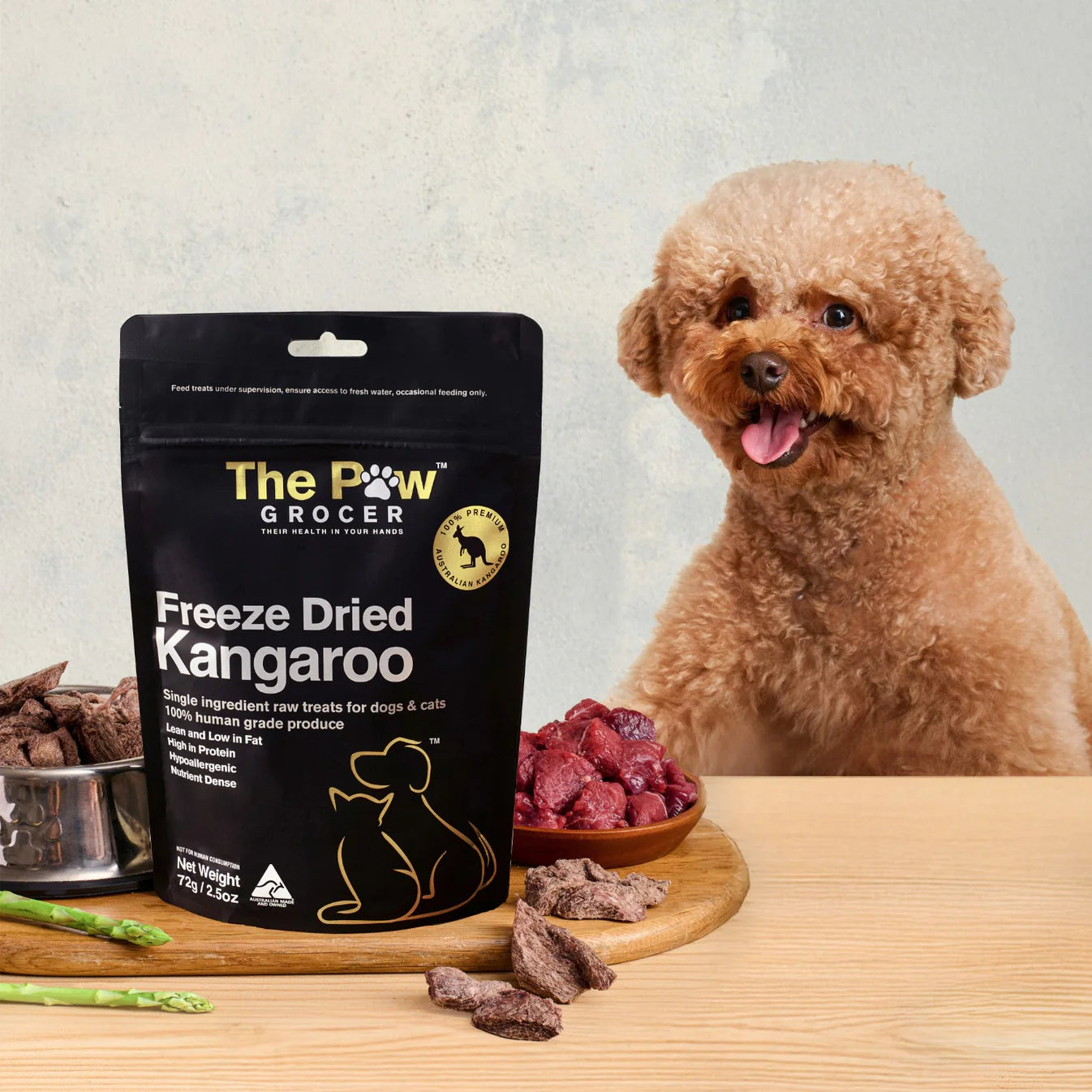 A happy poodle dog eagerly waiting for a high-value reward of The Paw Grocer Freeze-Dried Kangaroo treats, displayed next to fresh raw meat and the black product pouch.