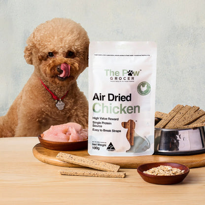 A fluffy poodle licking its lips next to a bag of The Paw Grocer Air Dried Chicken. Features raw chicken, oats, and a bowl full of easy-to-break chicken straps.
