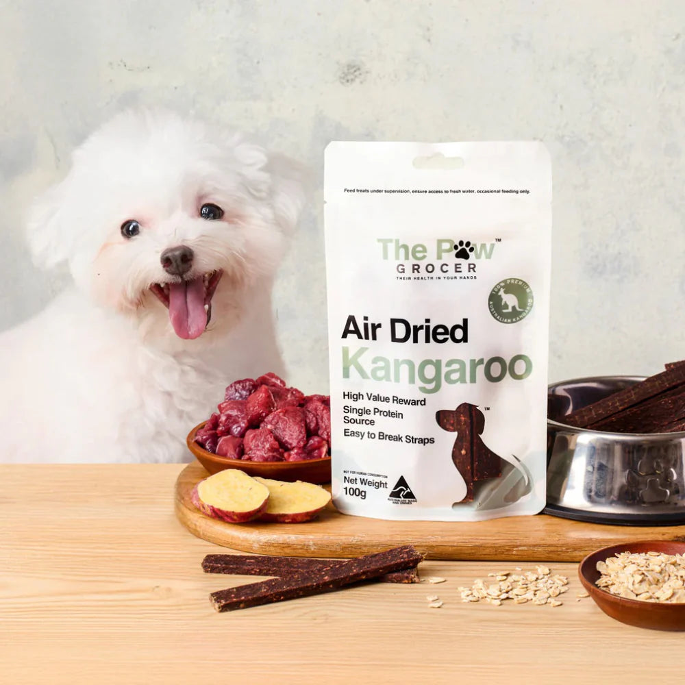 A happy white Bichon Frise dog waiting for The Paw Grocer Air Dried Kangaroo treats. Features fresh raw kangaroo, sweet potato, and a bowl of dried straps.