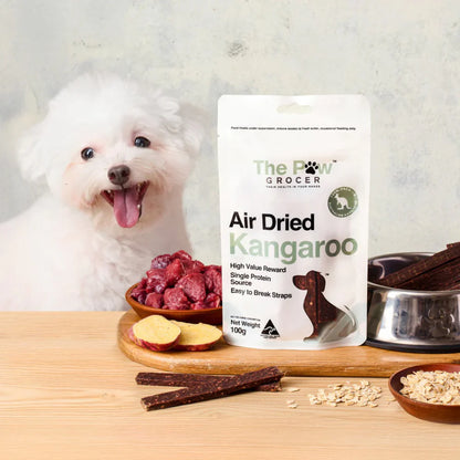 A happy white Bichon Frise dog waiting for The Paw Grocer Air Dried Kangaroo treats. Features fresh raw kangaroo, sweet potato, and a bowl of dried straps.