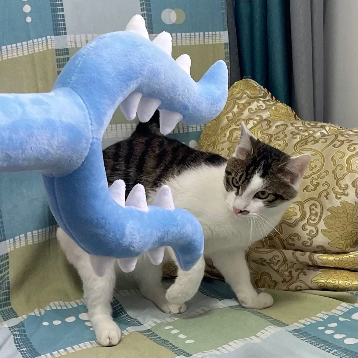 A playful tabby cat on a bed staring intensely at an approaching oversized blue plush defender fork toy, ready to pounce and unleash its hunting instincts.
