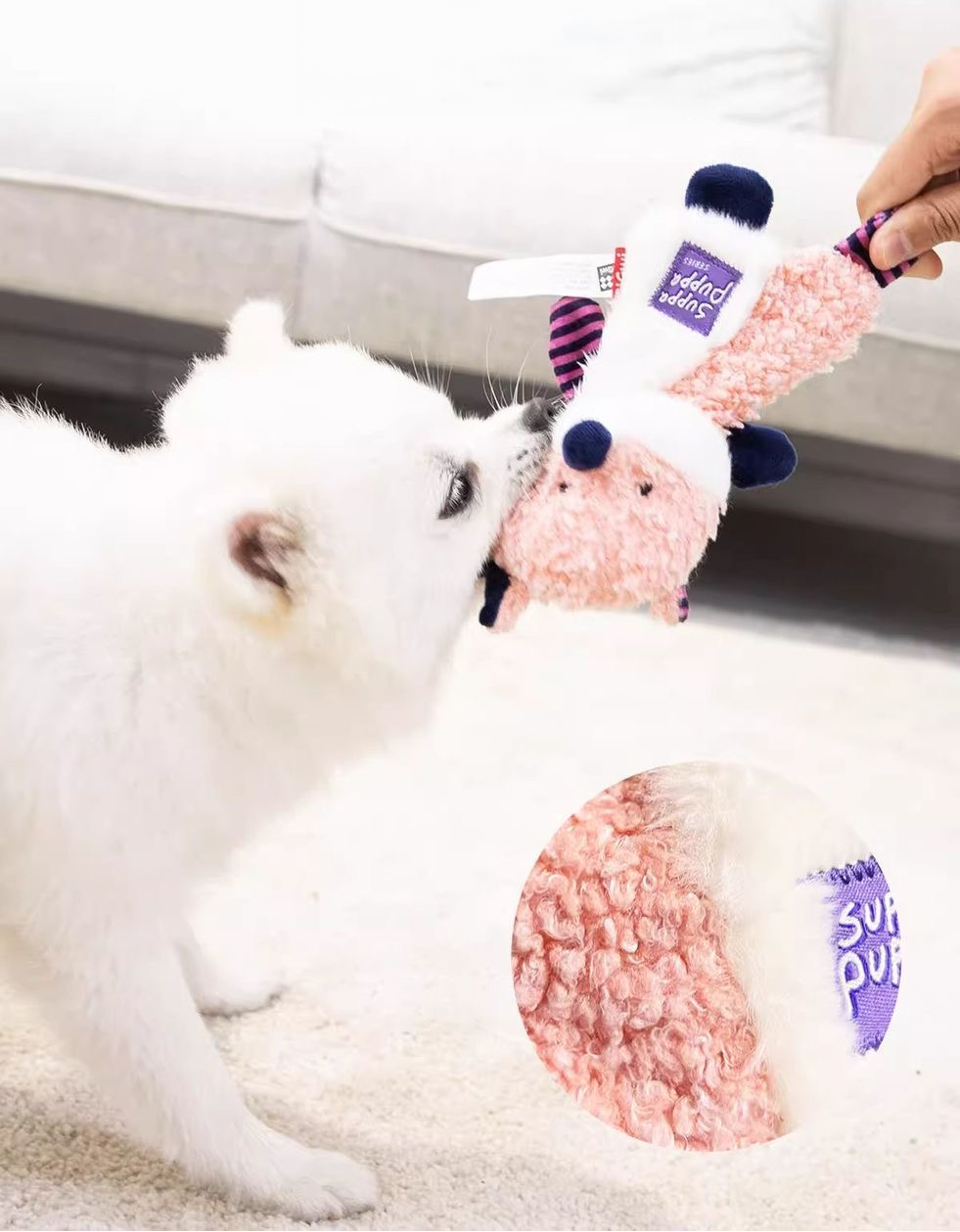 A white dog playfully pulling on the pink and white fox plush toy held by a person, featuring a circular inset that highlights the premium, scent-absorbing fleece texture.