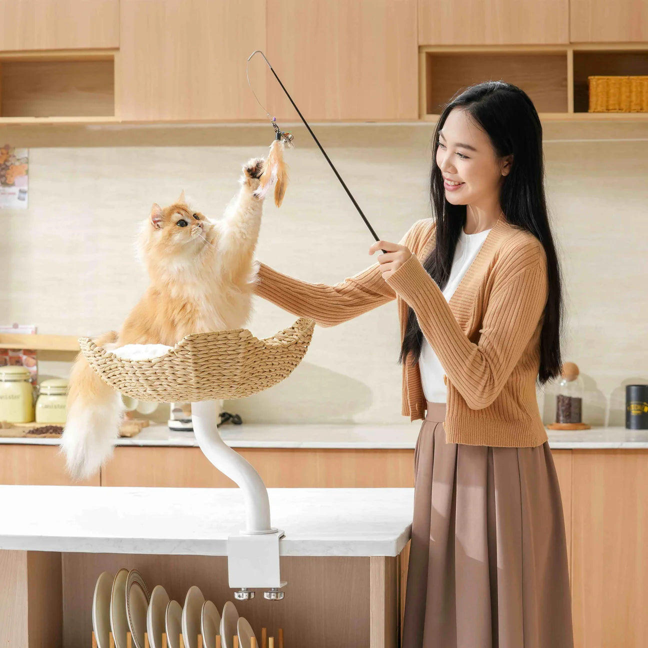 A woman smiling and playing with her golden cat using a feather wand. The cat is sitting securely in the white Michu DeskMate attached to a kitchen island countertop.