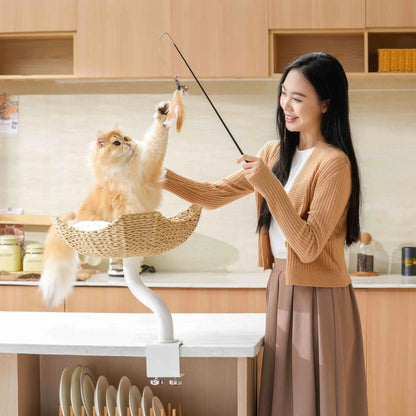 A woman smiling and playing with her golden cat using a feather wand. The cat is sitting securely in the white Michu DeskMate attached to a kitchen island countertop.
