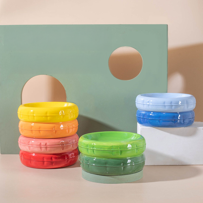 A stylish display of several stacked two-tone ceramic cat bowls in yellow, pink, green, and blue, arranged against a modern geometric backdrop.