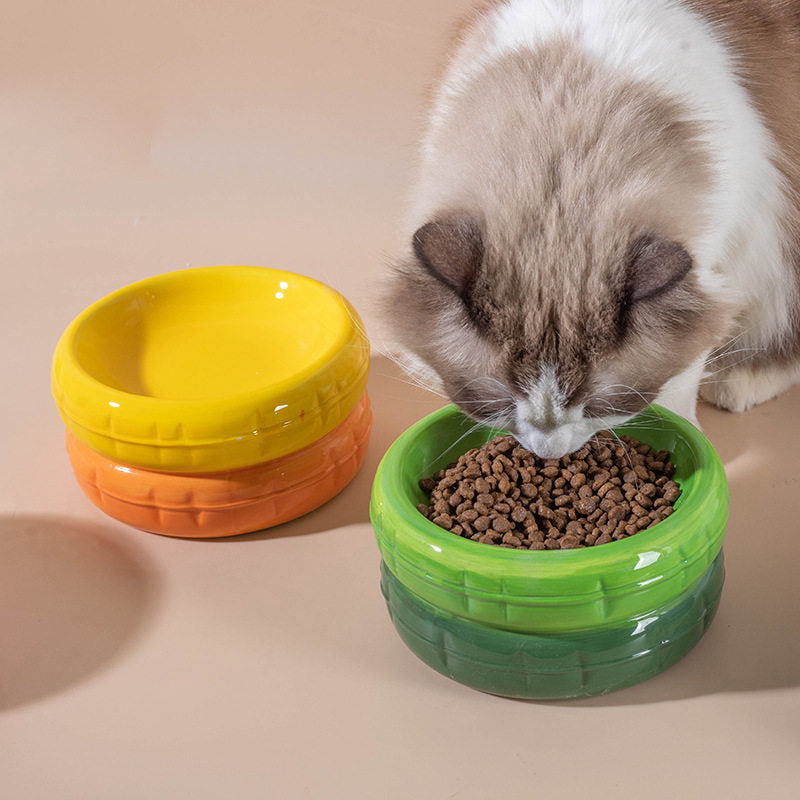 A fluffy cat eating dry food from a vibrant two-tone green ceramic cat bowl, with a yellow and orange bowl placed beside it on a warm neutral surface.