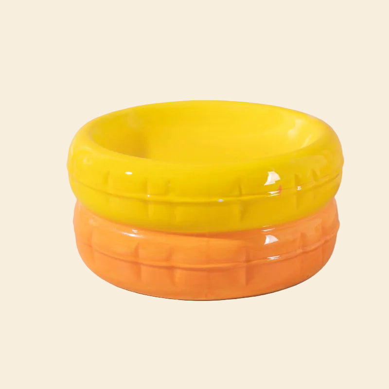 A vibrant two-tone ceramic cat bowl featuring a bright yellow top and a warm orange base, with a textured tire-like grooved pattern around the sides and a glossy finish.