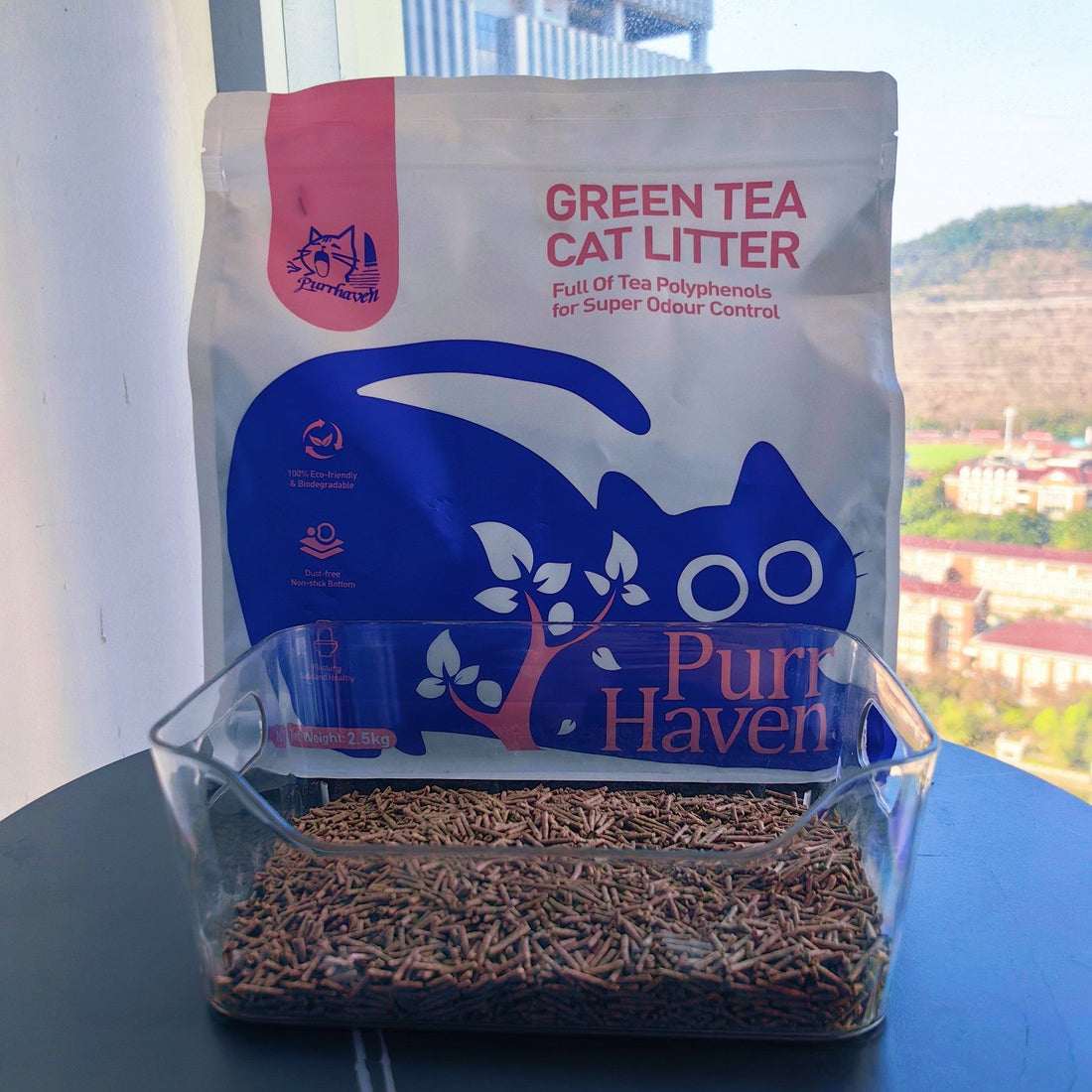 A bag of PurrHaven Green Tea Cat Litter placed behind a clear plastic litter box filled with the green tea tofu litter pellets, set against a bright window background with a city view.