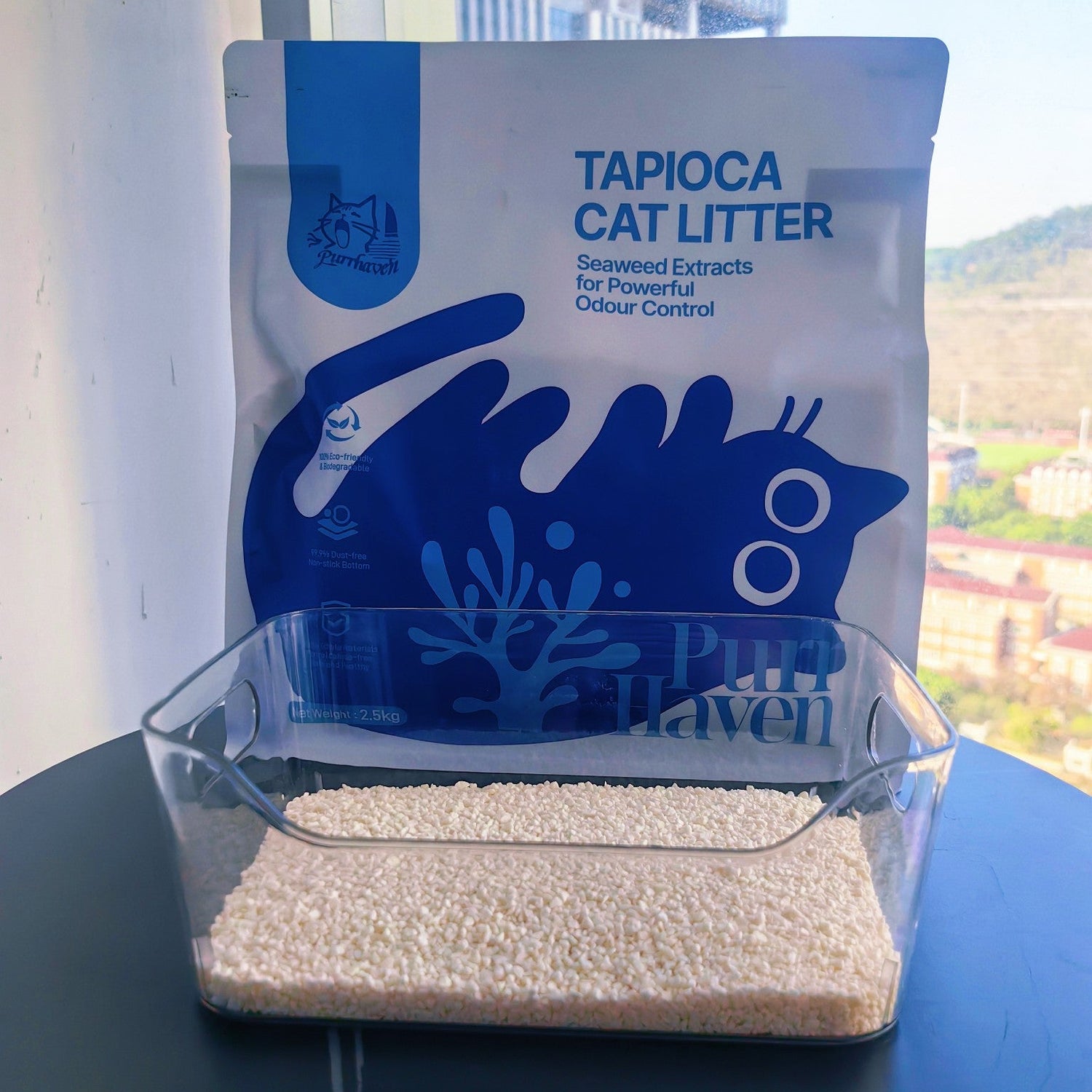A 2.5kg bag of PurrHaven Tapioca Cat Litter positioned behind a clear litter box filled with white, natural tapioca litter pellets, set against a bright window background with a city view.