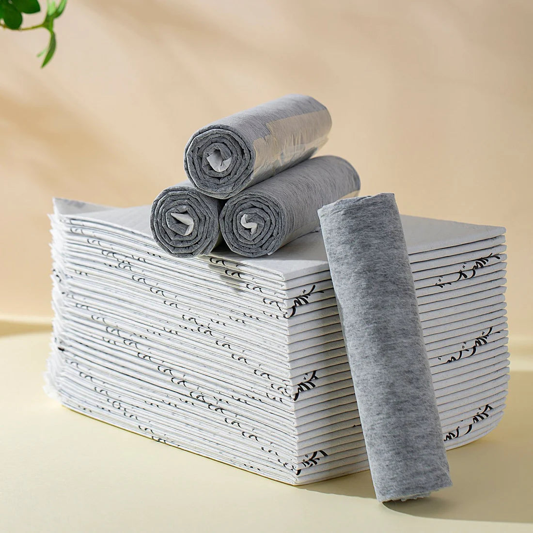 A neatly stacked pile of LYLLI PILLY puppy pads featuring a baby-grade comfort surface, with rolled-up grey pads against a warm beige background.