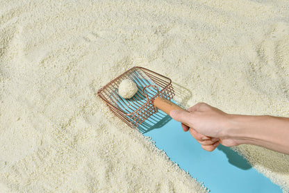Hand holding a small scoop with a ball of white clay on a sandy surface.