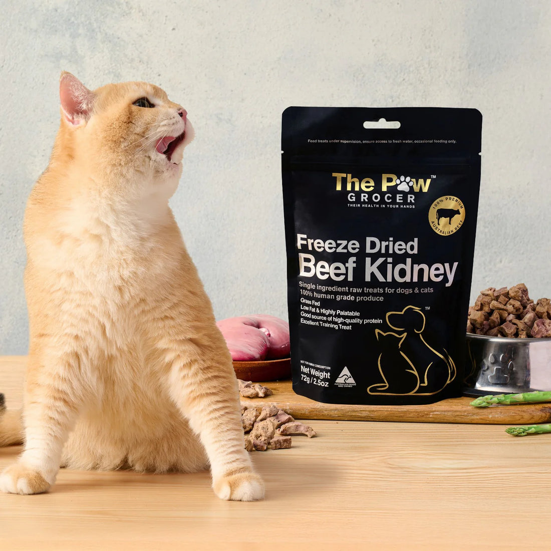 An adorable orange cat licking its lips eagerly next to The Paw Grocer Freeze-Dried Beef Kidney, a highly palatable, grass-fed organ superfood treat displayed with fresh raw kidney.